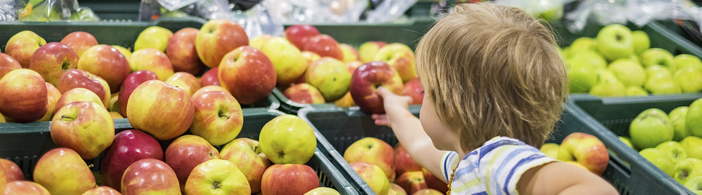 A young boy in the produce aisle of a grocery store, reaching for an apple.; Gettyimages: 960638210